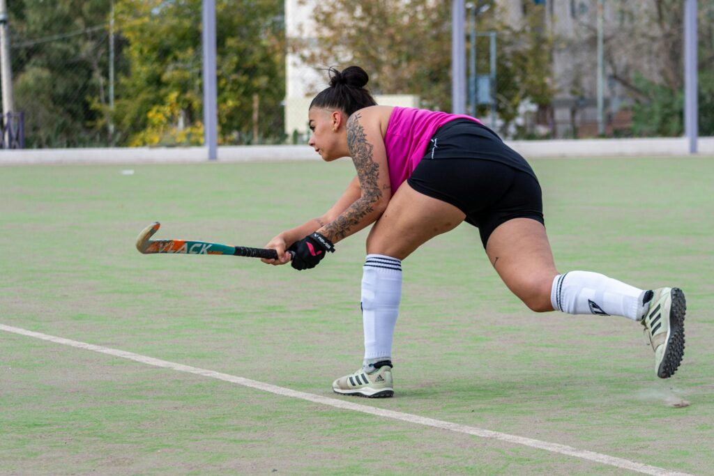 A female hockey player wearing a pink top playing field hockey on an outdoor pitch.